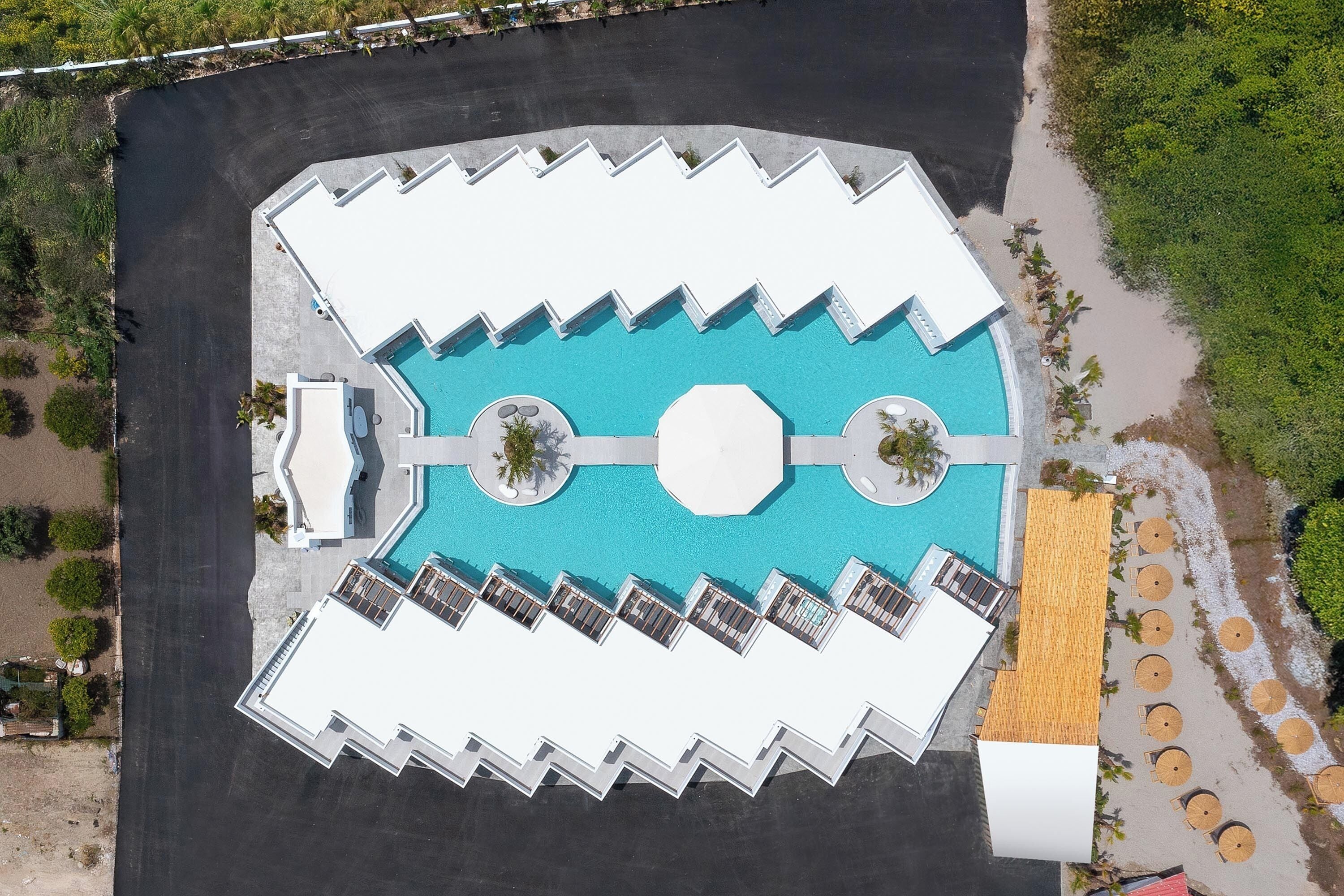 An aerial view of a resort with a central swimming pool, multiple lounge areas, and a beach area with umbrellas.