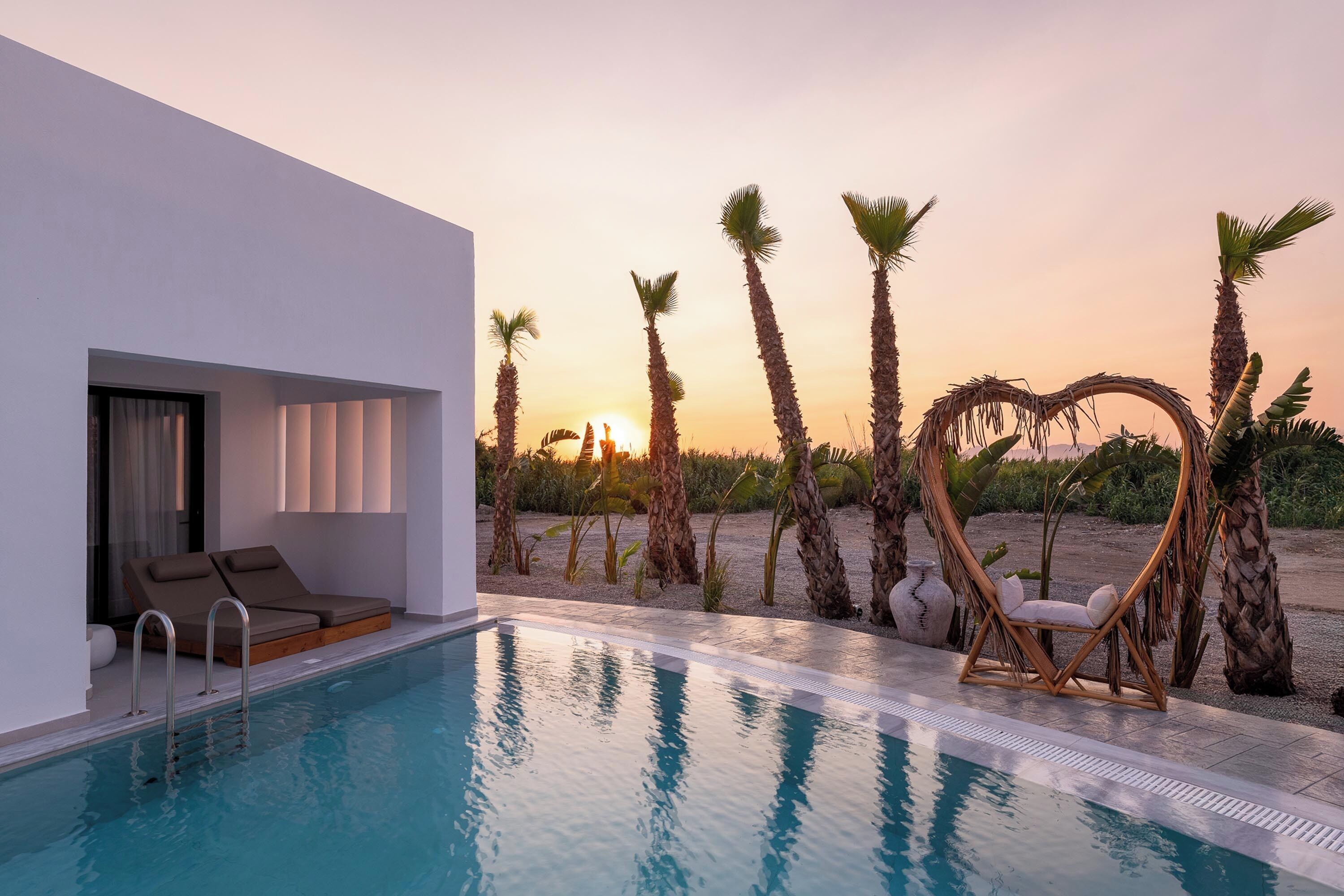 Un área de piscina moderna con sillones de descanso, un adorno con forma de corazón y palmeras al atardecer.