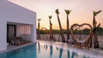 Un área de piscina moderna con sillones de descanso, un adorno con forma de corazón y palmeras al atardecer.