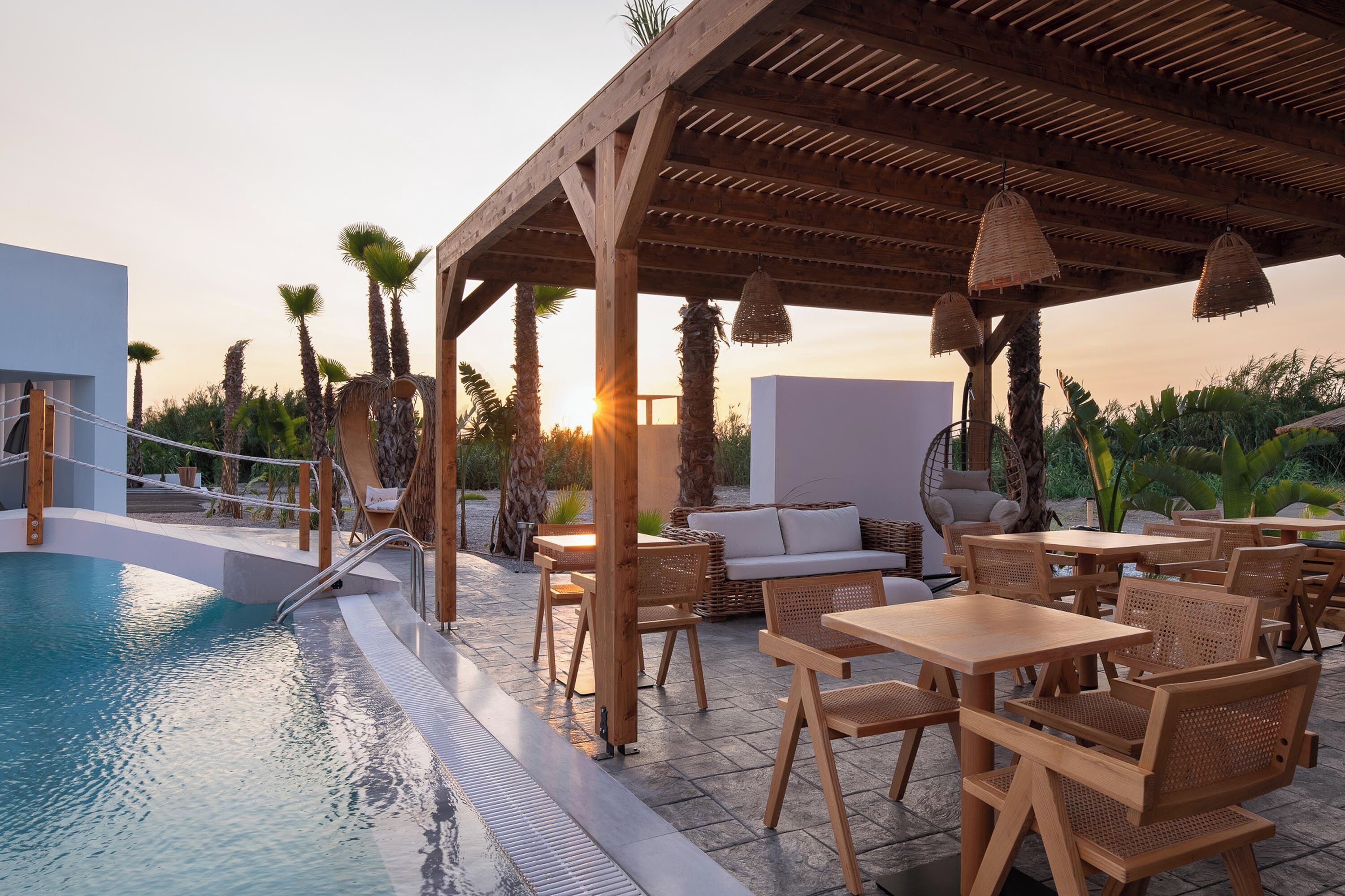 Un espace aménagé au bord de la piscine, avec des tables et des chaises en bois, un coin salon couvert et une vue sur le coucher du soleil.