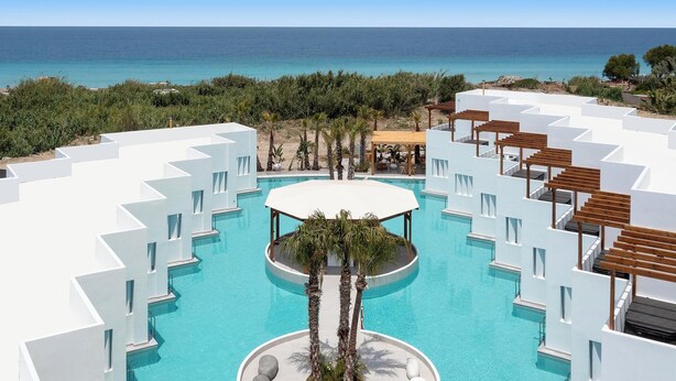 A resort with a central pool surrounded by white buildings, a beach view, and palm trees.