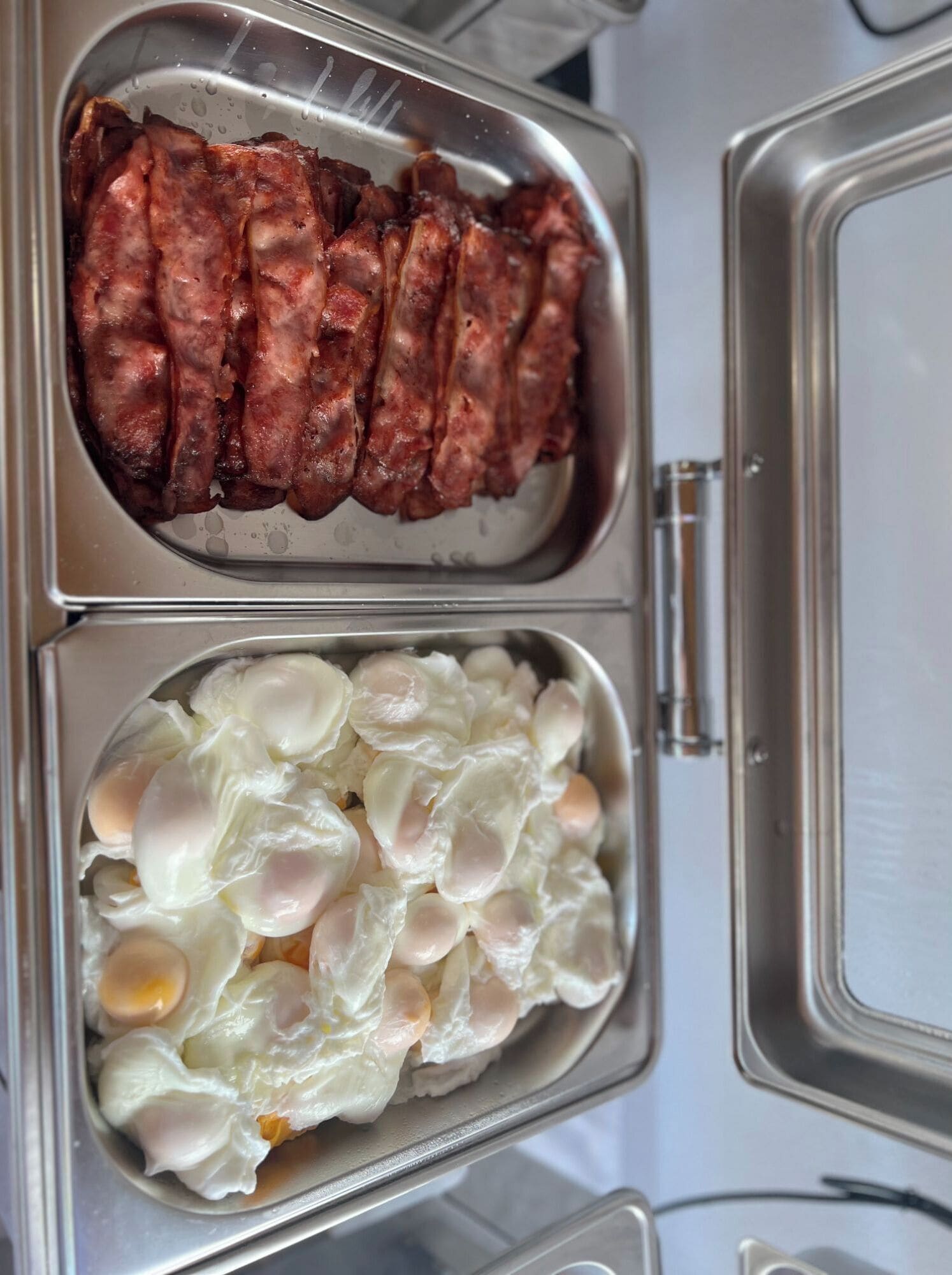 A kitchen with two stainless steel trays, one containing cooked bacon and the other containing poached eggs.