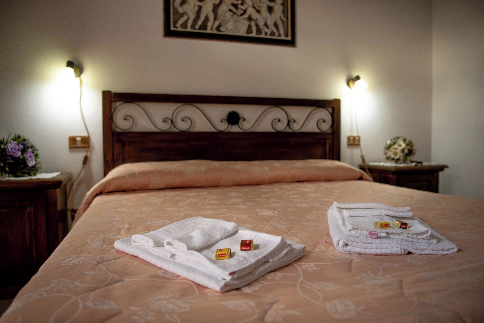 Una cama bien tendida con toallas dobladas y bombones, cabecera de madera y lámparas de pared.