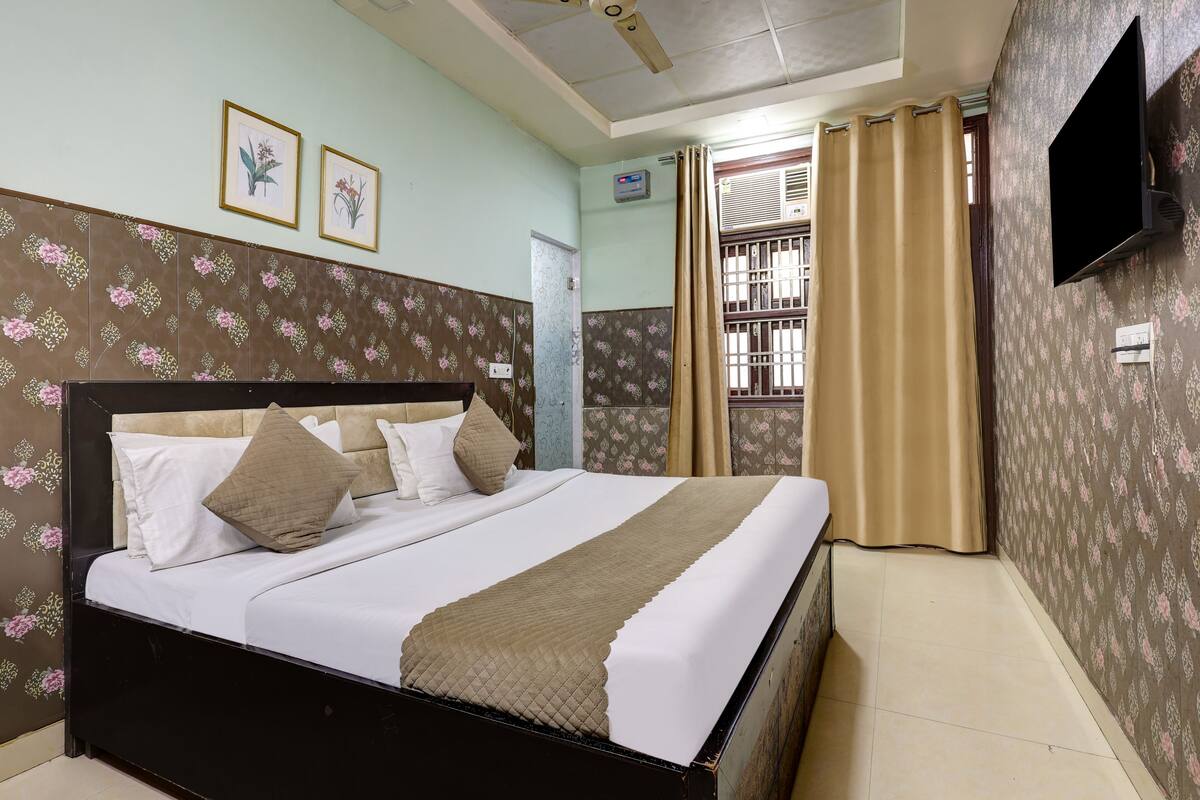 A neatly made bed with a headboard, a flat-screen TV mounted on the wall, and a window with curtains.