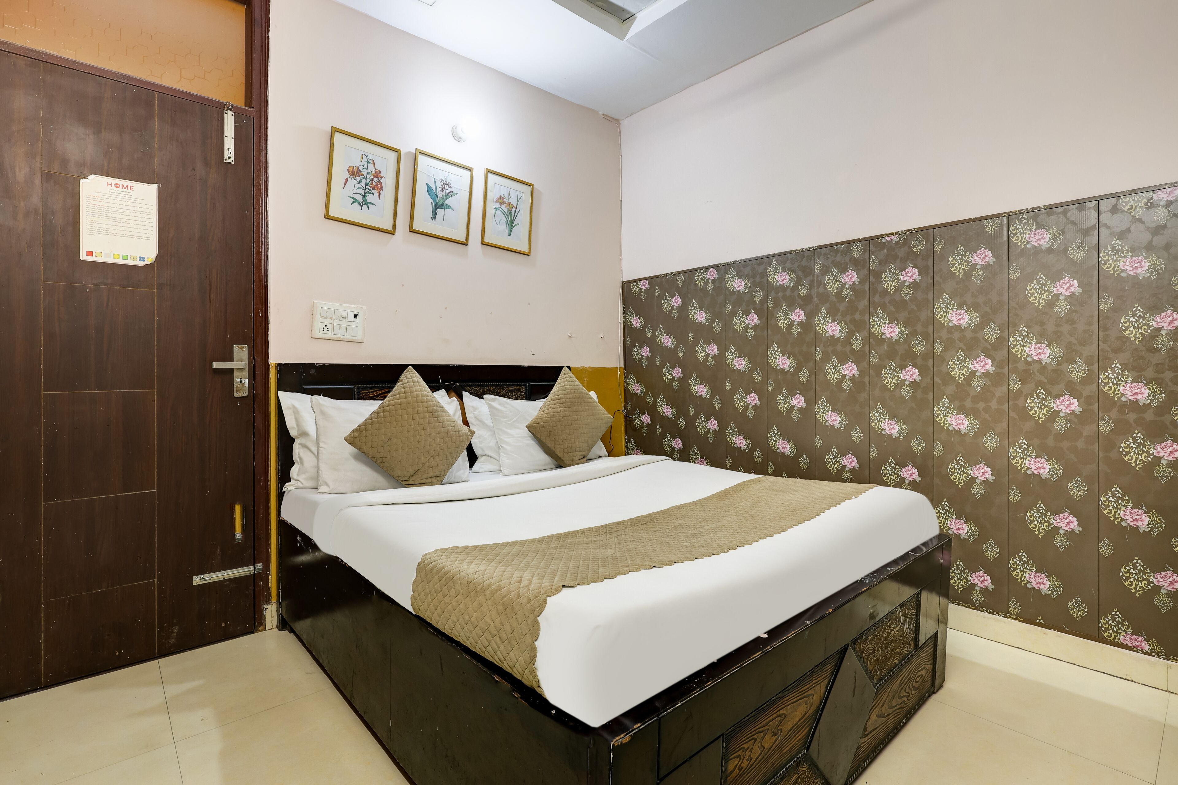 A neatly made bed with a white and beige bedspread, a wooden headboard, and a brown wardrobe with a floral pattern. There are three framed pictures on the wall above the bed.