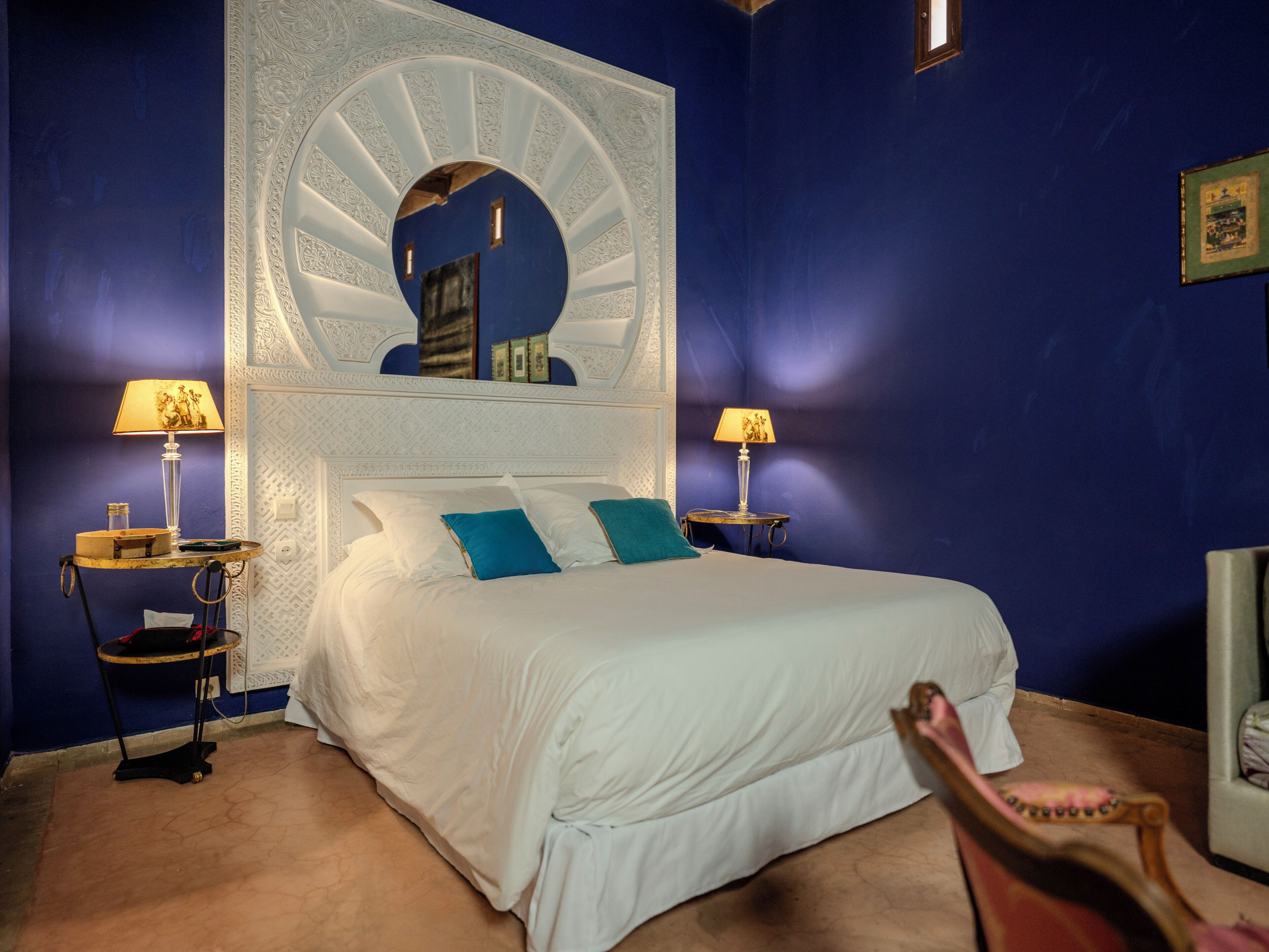 A bedroom with a large bed, two bedside tables with lamps, a mirror above the bed, and a chair in the corner.
