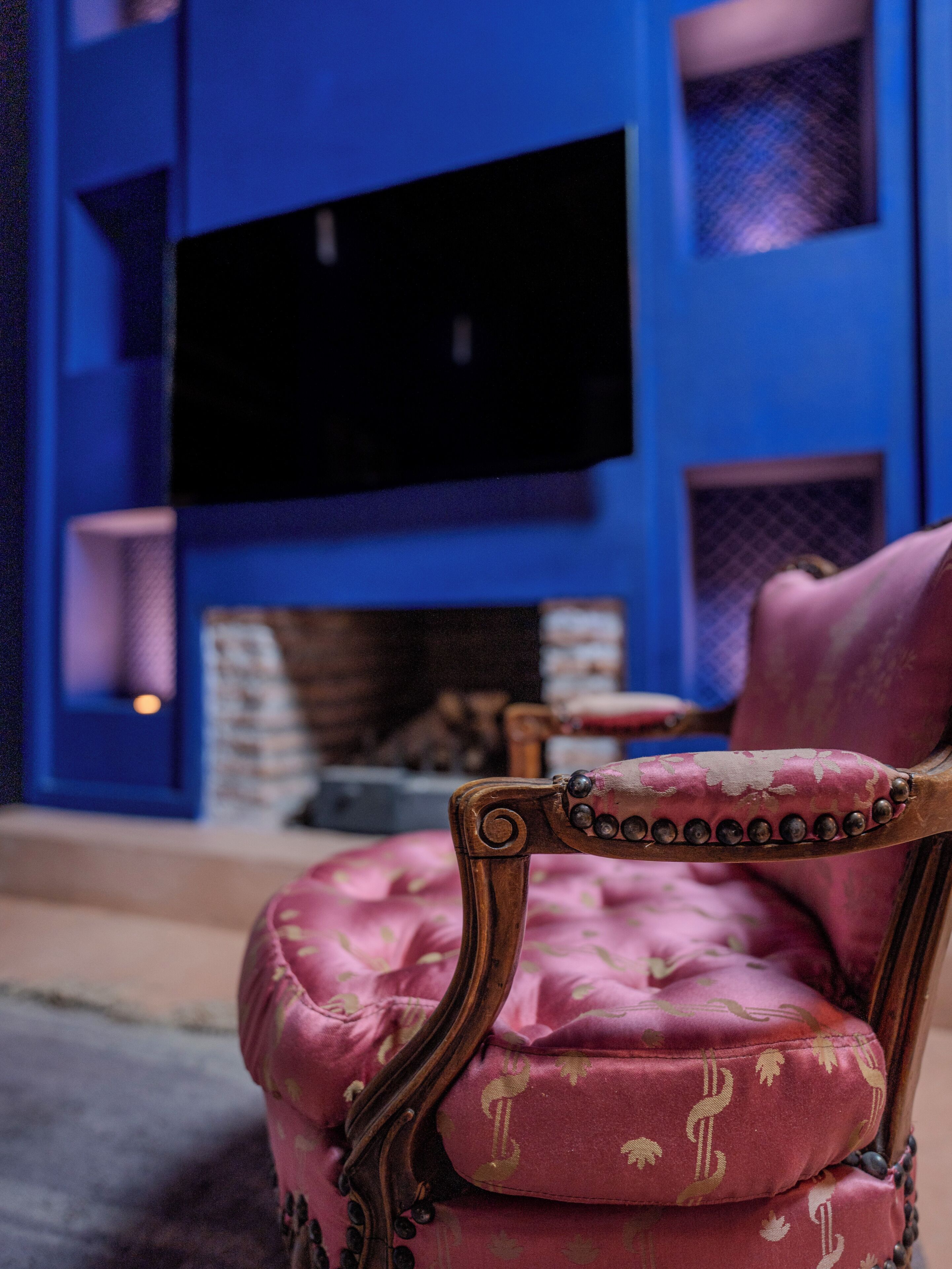 A plush pink armchair with a decorative backrest in a room with a blue accent wall and a flat-screen TV mounted above a fireplace.