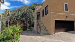 Ein zweistöckiges Haus mit einer Steintreppe, die zu einem Balkon führt, umgeben von üppigem Grün und blauem Himmel.