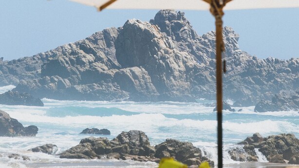 Ein Strand mit einem weißen Sonnenschirm, felsigen Klippen und Wellen im Hintergrund.