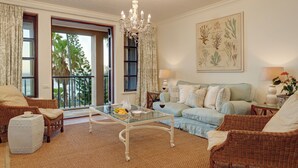 Ein geräumiges Wohnzimmer mit einem Glastisch, Korbmöbeln, einer Chandelierleuchtre und einem Balkon mit Blick auf Palmen.