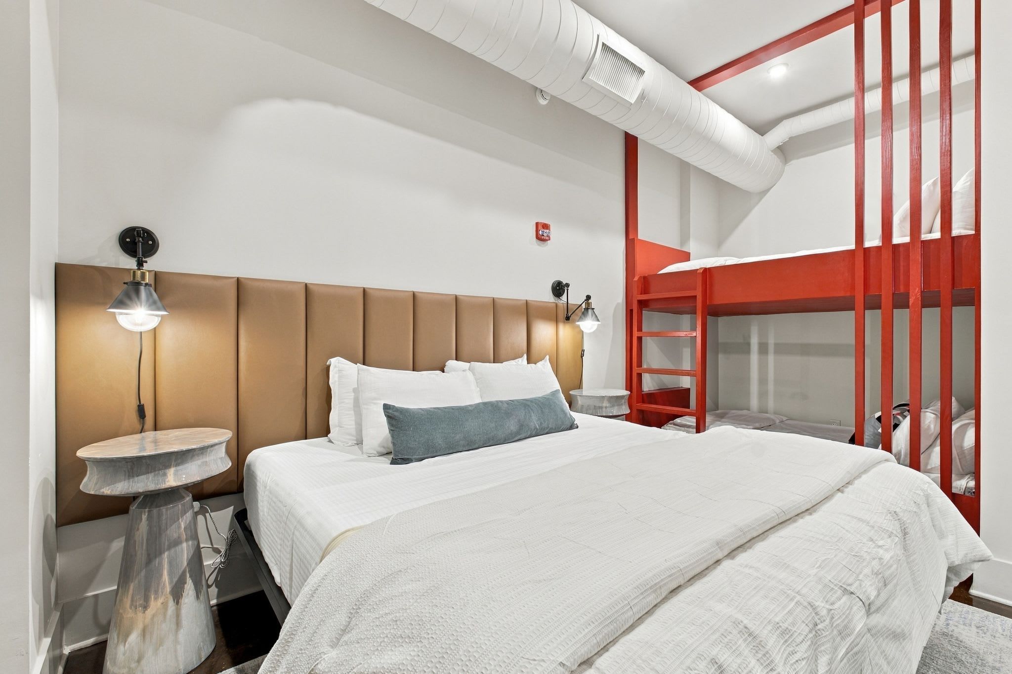 Camera d'albergo moderna con un letto grande, un tavolino rotondo, una cuccetta rossa e una bocchetta di ventilazione al soffitto.