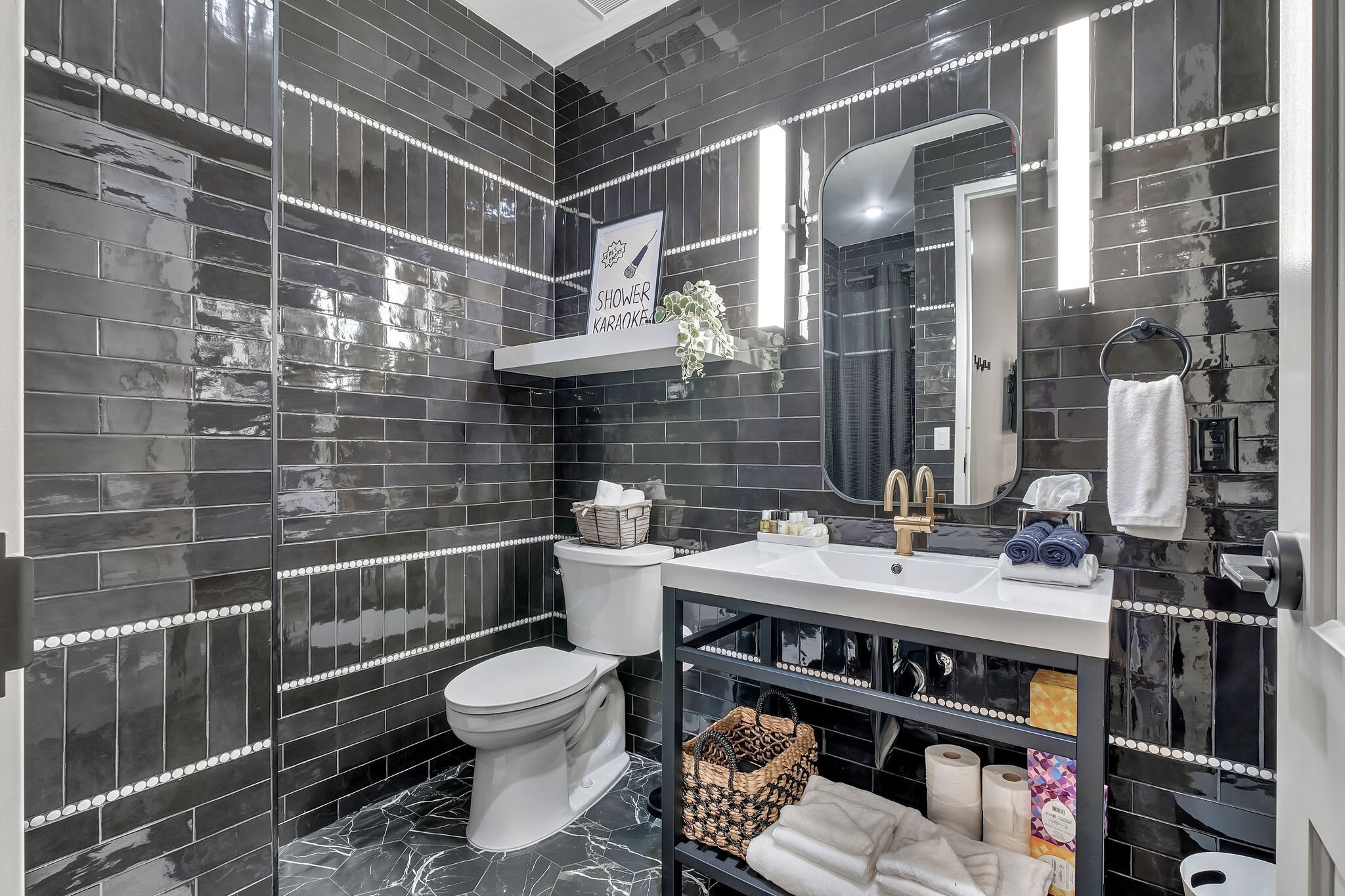 A modern bathroom with black tiled walls, a white toilet, a vanity with a mirror, and a shower area.