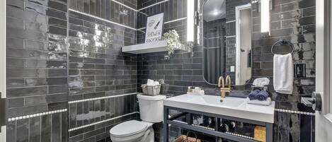 A modern bathroom with black tiled walls, a white toilet, a vanity with a mirror, and a shower area.