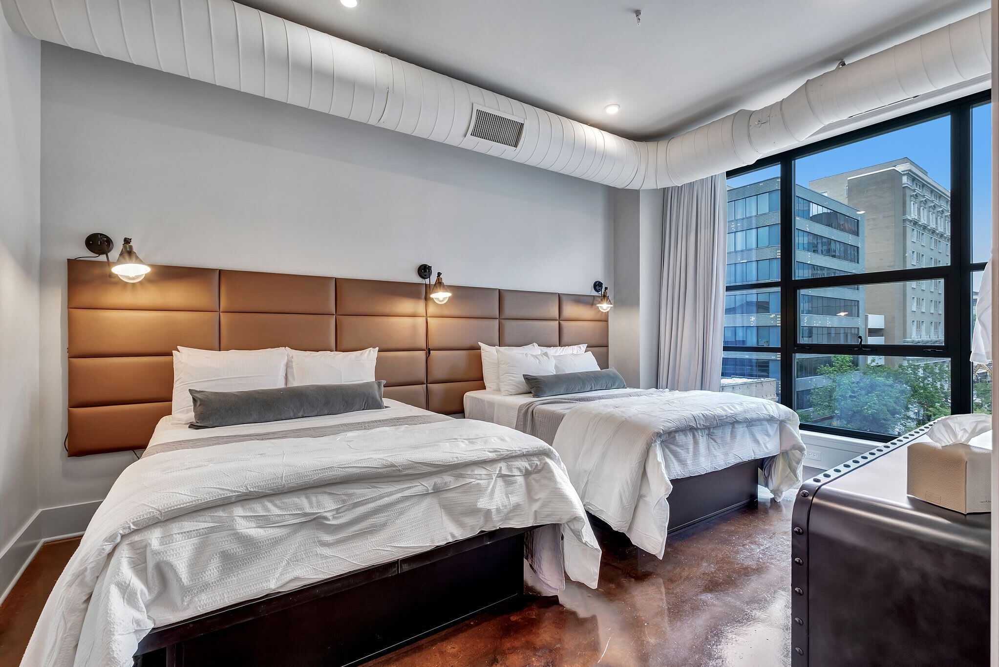 Une chambre d’hôtel moderne avec deux lits, une grande fenêtre donnant sur la ville et un plafond blanc équipé de conduits de ventilation.