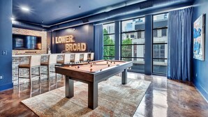 Una mesa de billar en una habitación con pared de ladrillo, zona de bar y ventanas amplias.