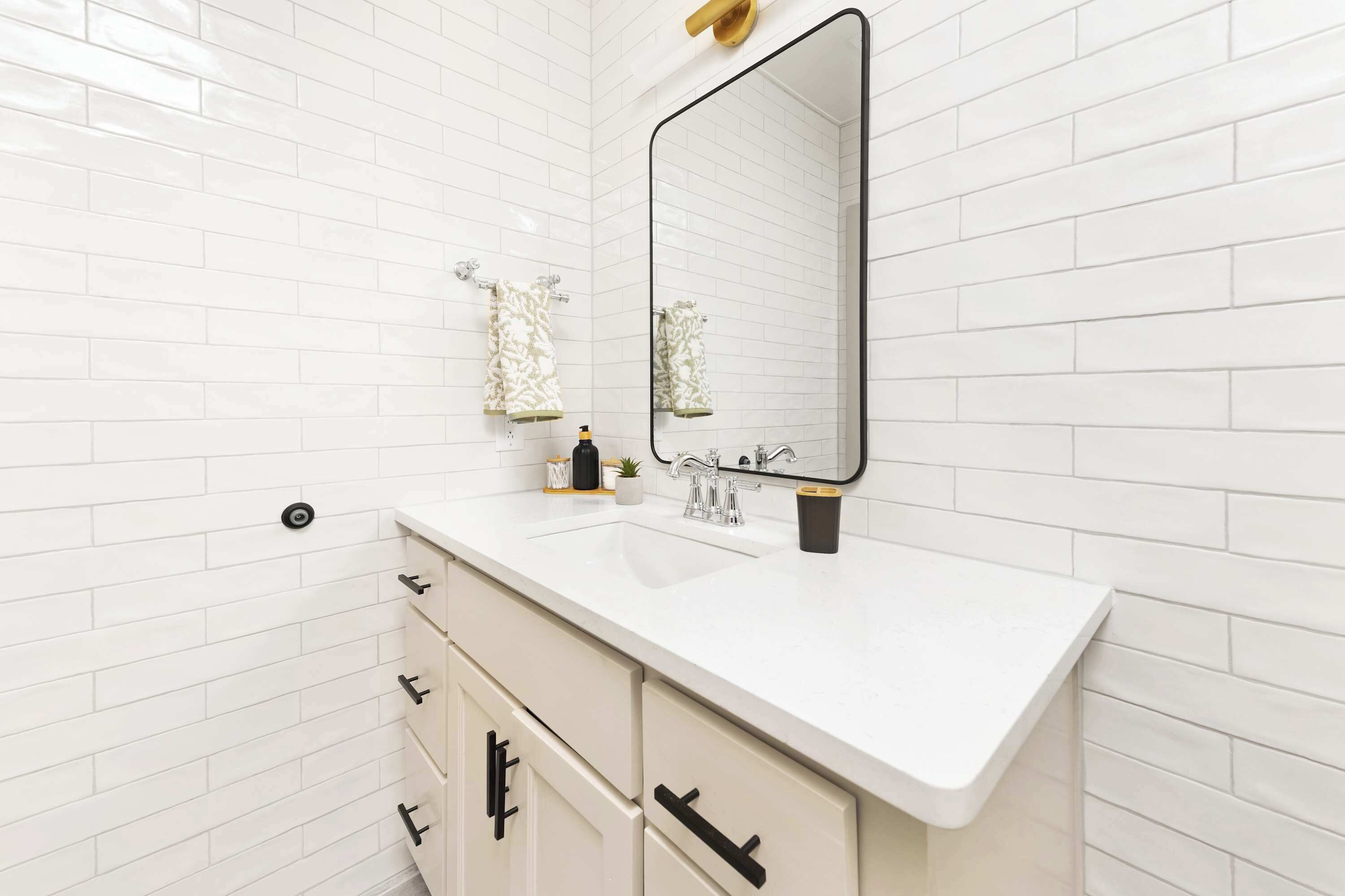 Une salle de bain avec des murs recouverts de carreaux blancs, un lavabo blanc avec un robinet noir et un miroir au-dessus du lavabo.