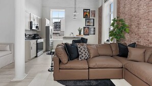 Una sala de estar moderna con un sofá seccional marrón, una cocina blanca con electrodomésticos de acero inoxidable y una pared de ladrillo.