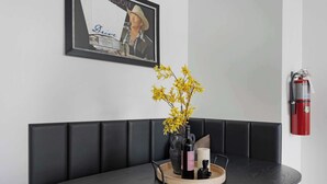 Un comedor moderno con un banco acolchado negro, una mesa de madera con una bandeja que sostiene una botella, un florero con flores amarillas y un cuadro en la pared.