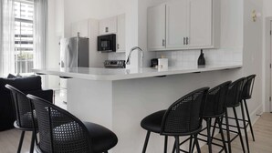 Una cocina moderna con gabinetes blancos, un refrigerador de acero inoxidable, un microondas y un fregadero. Hay cuatro taburetes negros y una ventana con cortinas.