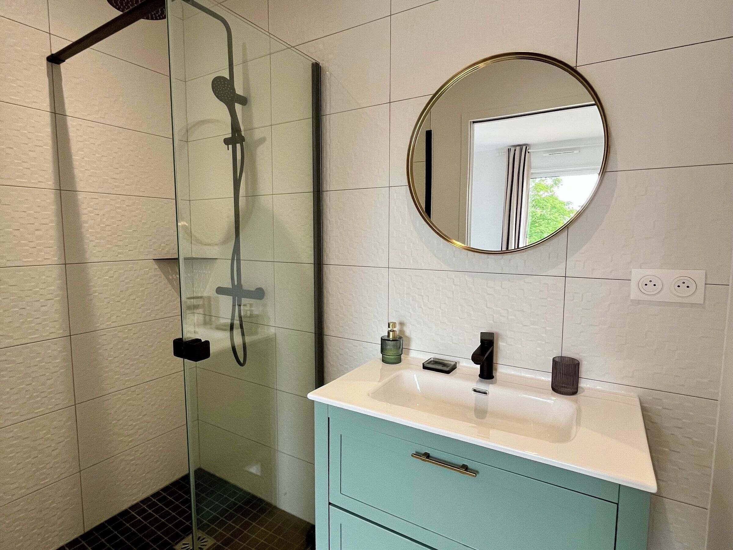 A bathroom with a glass-enclosed shower, a white vanity with a round mirror, and a teal cabinet.