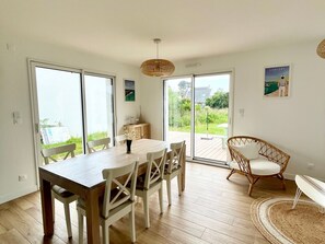 A dining area with a wooden table and chairs, a wicker chair, and a view through glass doors to a garden. - New and Spacious House Near Port Tudy (Groix)