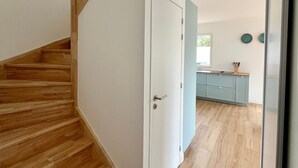 Una escalera conduce a una habitación con una puerta blanca, una zona de cocina con armarios azul claro y un ventanal con vistas a la vegetación.
