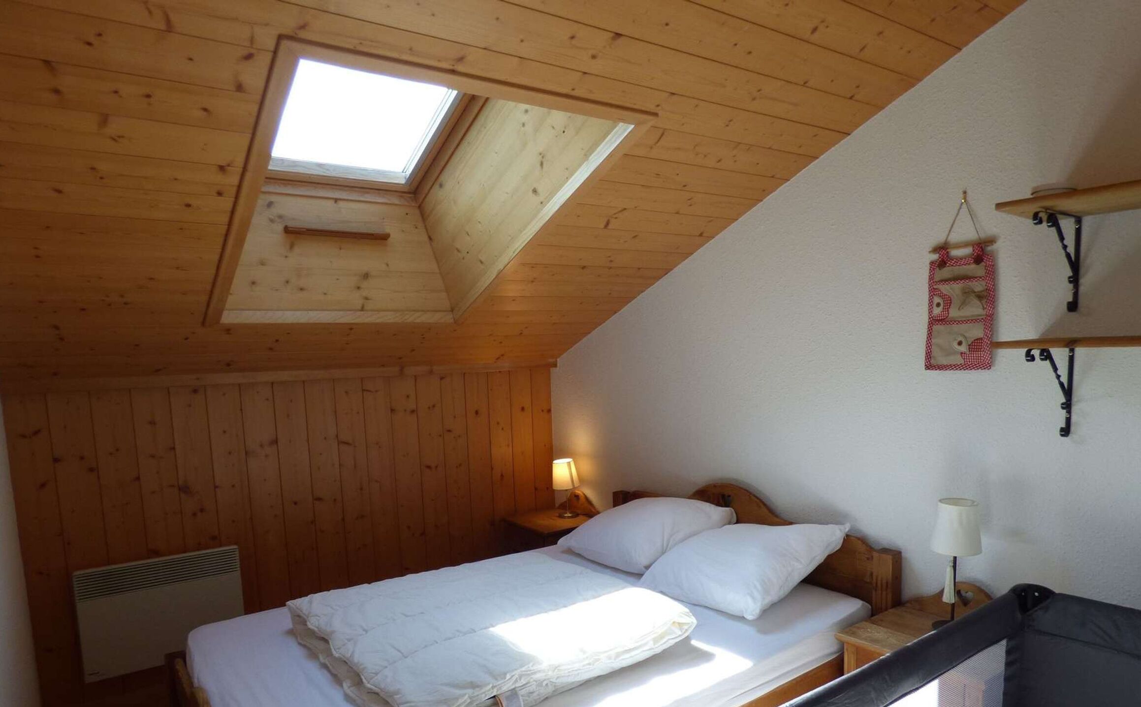 Un dormitorio con techo inclinado, tragaluz, cama con ropa de cama blanca, mesita de noche con lámpara y estante de pared con un objeto decorativo.