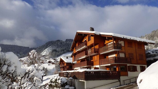 A wooden chalet with multiple balconies, surrounded by snow-covered trees and mountains.