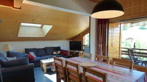 A cozy room with a sloped ceiling, a sofa, a TV, a dining table with chairs, and a window with curtains.