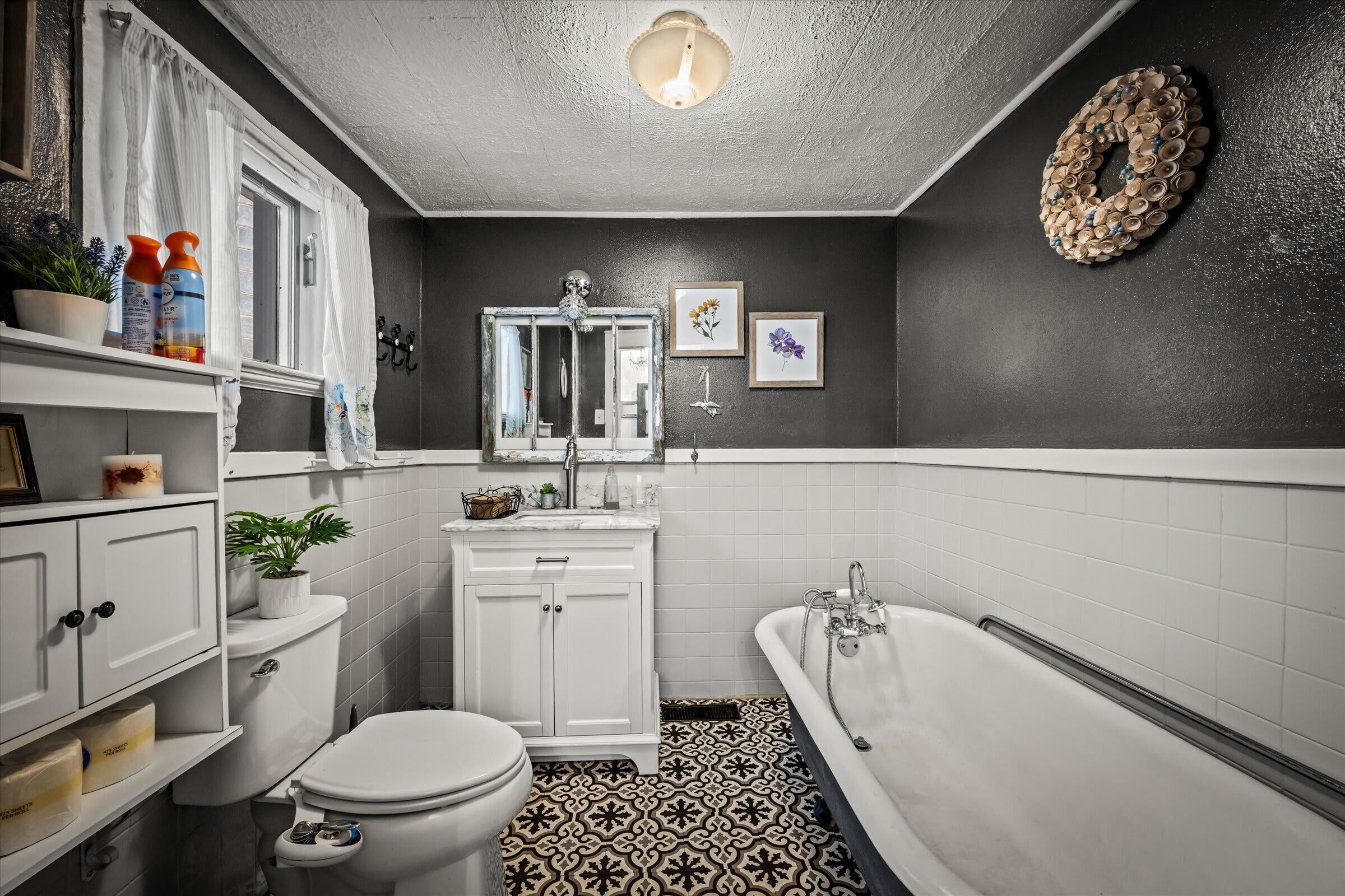 A bathroom with a toilet, sink, and bathtub. There are bottles on the shelf, a mirror, and framed pictures on the wall.