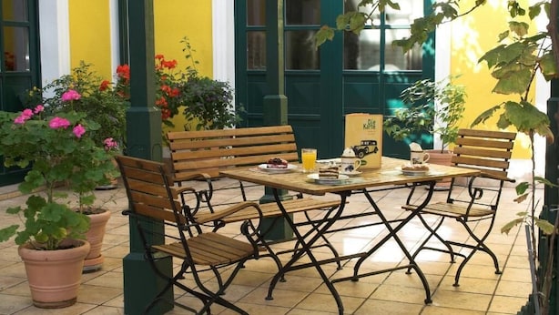 Un café al aire libre con bancos de madera y una mesa, rodeado de macetas con plantas y una pared amarilla.