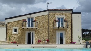 Un edificio de dos plantas con fachada de piedra, piscina delantera y balcones con jardineras florales.