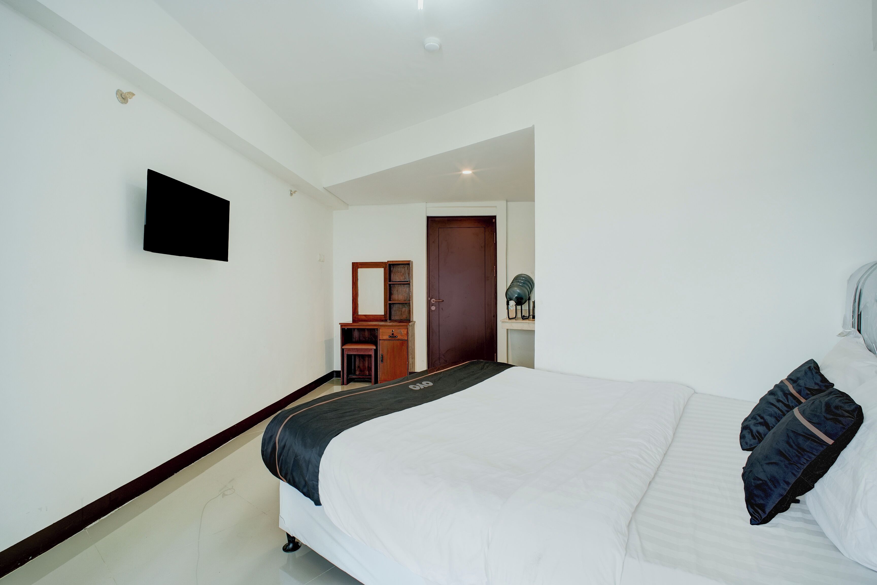 A bedroom with a bed, a flat-screen TV mounted on the wall, a wooden dresser, and a door leading to another room.