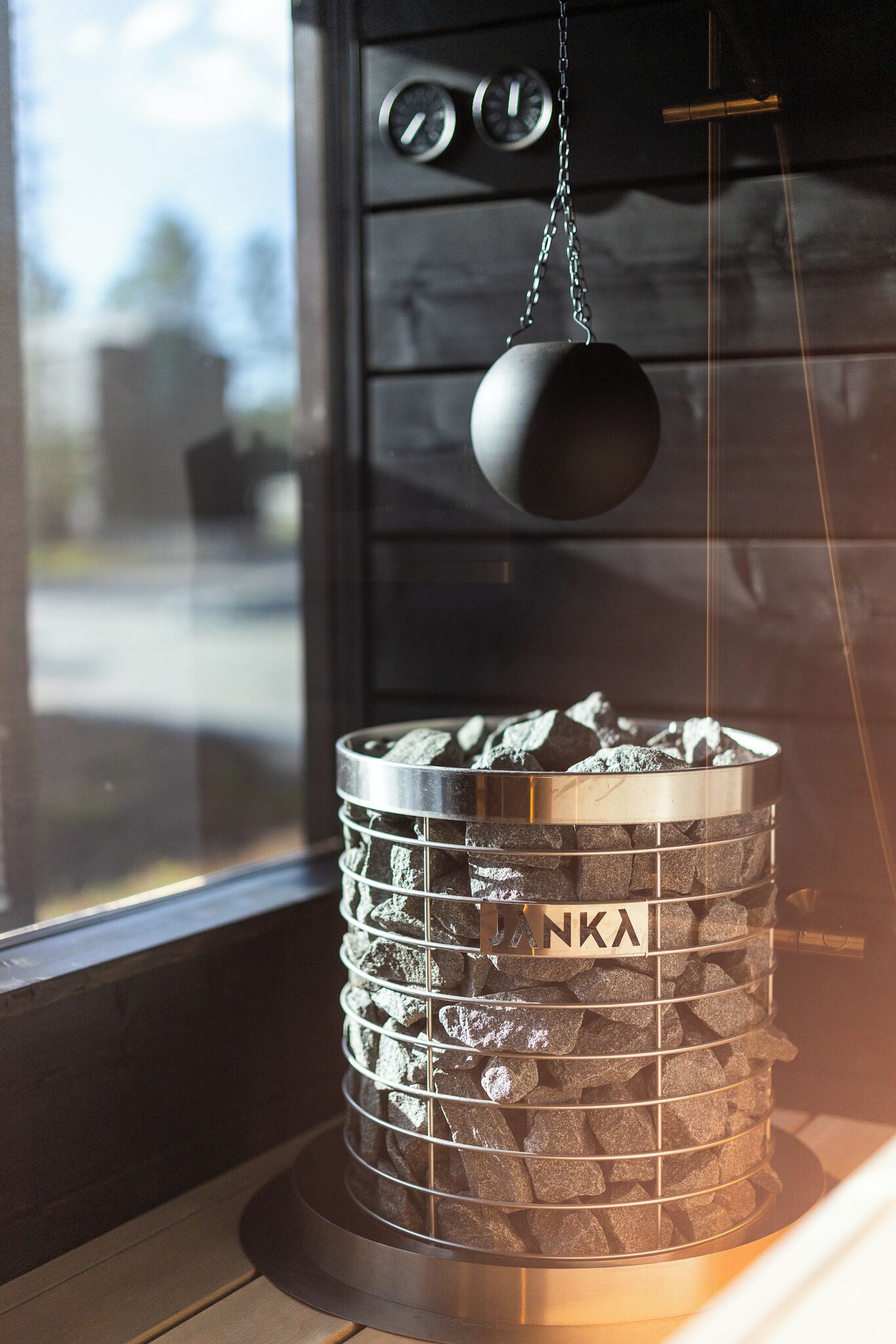 A cylindrical charcoal grill with a metal mesh and a hanging black weight, labeled 'ANKA,' against a dark wooden backdrop.