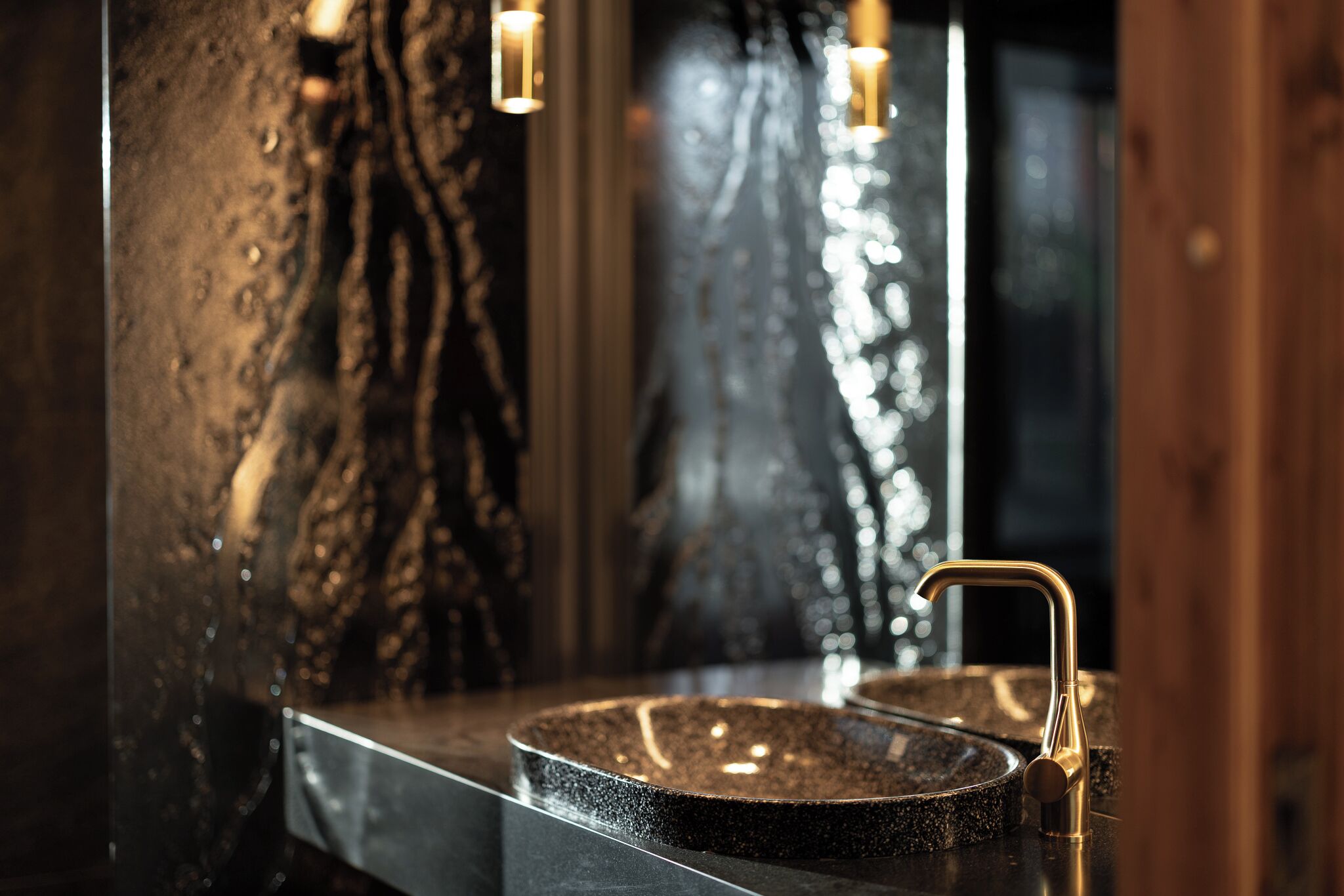 Un bagno con un lavabo in granito, un rubinetto dorato e uno specchio che riflette una parete strutturata.