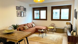 Una sala de estar con un sofá, una mesa de comedor con sillas, una mesa de centro y una lámpara. Hay ventanas con persianas, una planta y cuadros en la pared.