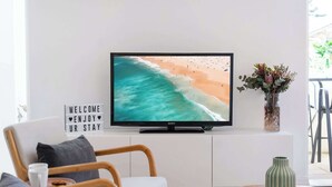 Un televisor sobre un mueble blanco, un cartel que dice 'WELCOME ENJOY UR STAY', una silla de madera, una mesita con un florero verde y una ventana con vistas a una playa.