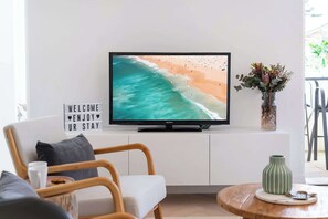 Une télévision posée sur un meuble blanc, une pancarte indiquant « BIENVENUE, PASSEZ UN BON SÉJOUR », une chaise en bois, une petite table avec un vase vert, et une fenêtre donnant sur une plage.