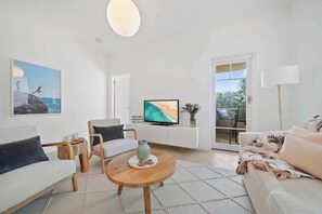 Un salon moderne avec un canapé, un fauteuil, une table basse, un téléviseur et une vue sur l’extérieur, visible à travers une porte vitrée.