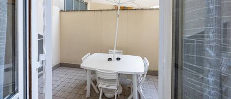 A small, enclosed patio area with a white table and chairs, a white umbrella, and a view of a building in the background.