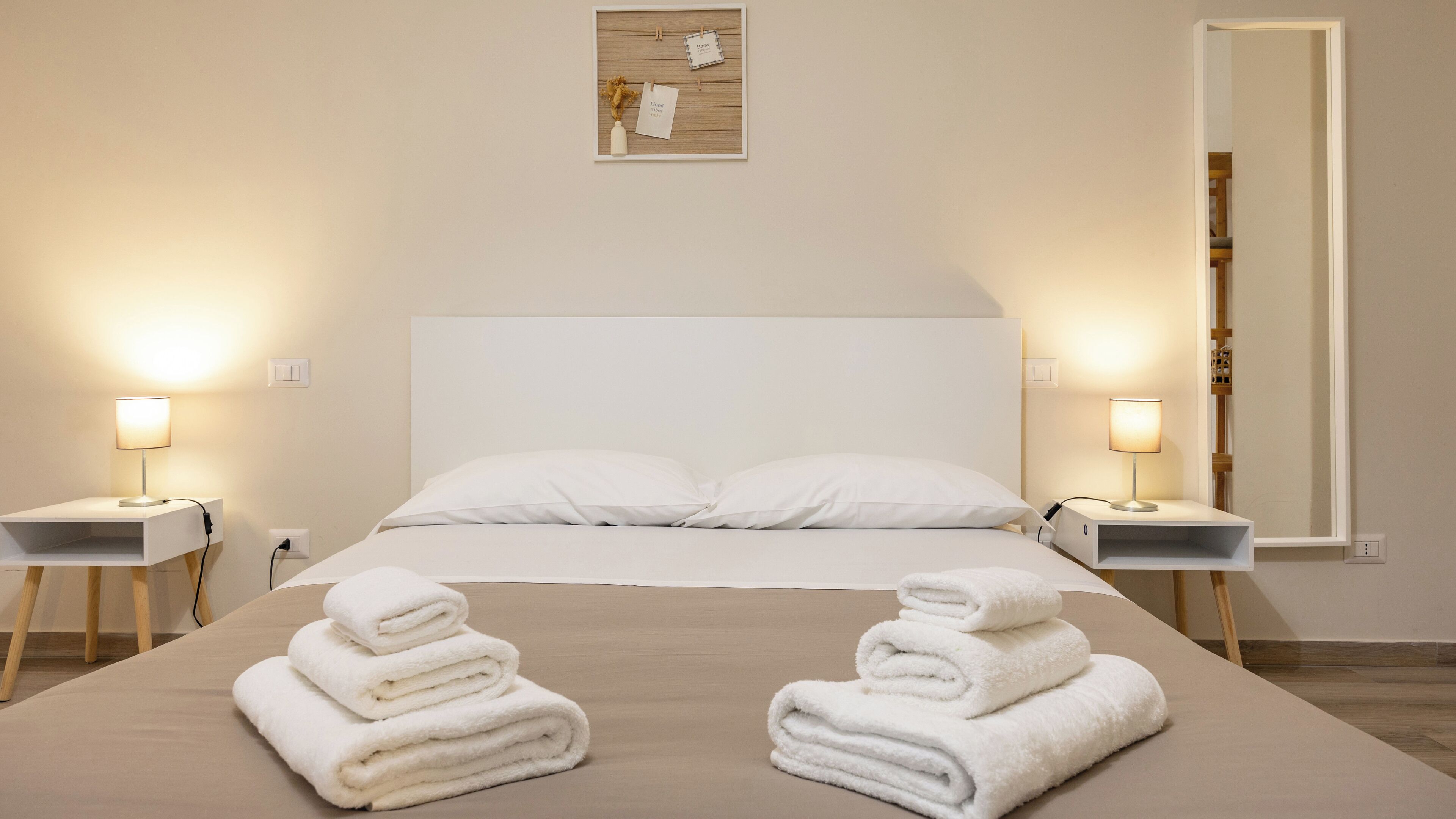 A neatly made bed with white linens and two folded towels on the bed. There are two bedside tables with lamps and a small framed picture on the wall.