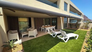 Een modern terras buiten met een houten tafel en witte stoelen, omgeven door kunstgras en witte loungestoelen.