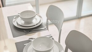 A dining table with a set of white dishes and cutlery, a chair, and a window in the background. - Astrum by Kozystay - Ciumbuleuit (Bandung)