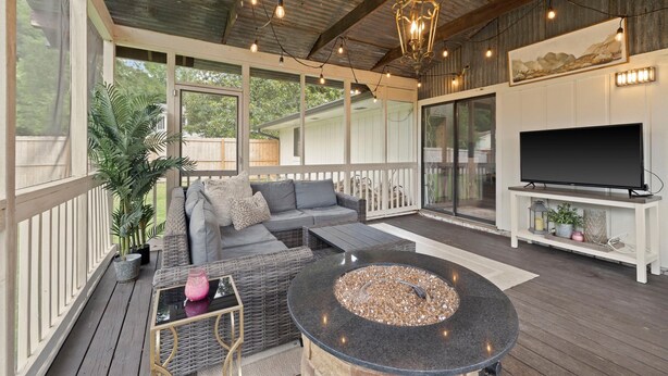 A covered outdoor living area with a sofa, a round table with a fire pit, a TV on a stand, and string lights.