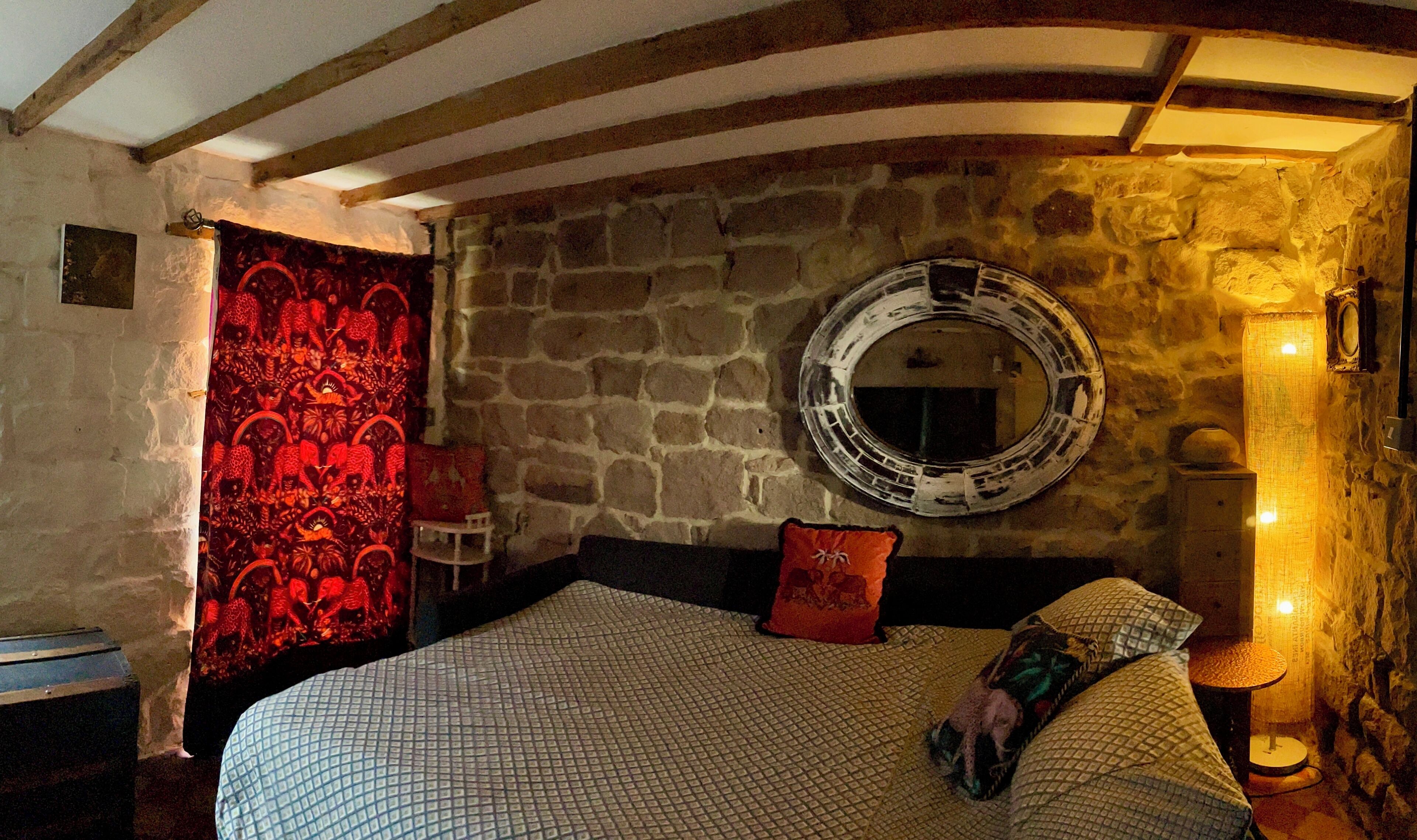 A bedroom with a stone wall, a round mirror, a bed with patterned bedding, and a red tapestry.