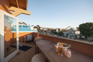 Ein Balkon mit Blick auf eine Wohngegend, ein Holztisch mit einem Getränk, eine Obstschale und ein Stuhl.