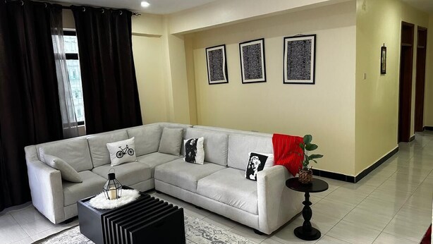 A modern living room with a grey sectional sofa, a black coffee table, and a small side table. There are three framed artworks on the wall and a red throw on the sofa. - Girrafe Apartments (Dar es Salaam)