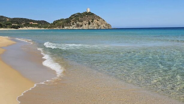 Una playa con agua cristalina, una orilla de arena y una colina con una estructura al fondo.