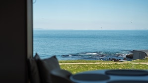 A view of the ocean from a room with a window, a small building, and a table with a circular object on it. - Peninsula Suites (Hellnar)
