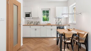 Eine moderne Küche mit weißen Schränken, einem Holzboden im Fischgrätmuster, einem Essbereich mit Holztisch und -stühlen und Blick ins Grüne.